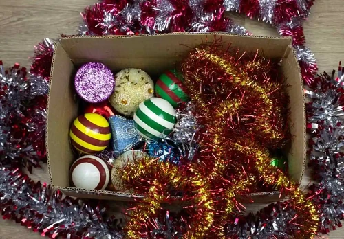 An open cardboard box filled with colorful Christmas ornaments and tinsel, showing mixed holiday decor stored loosely.