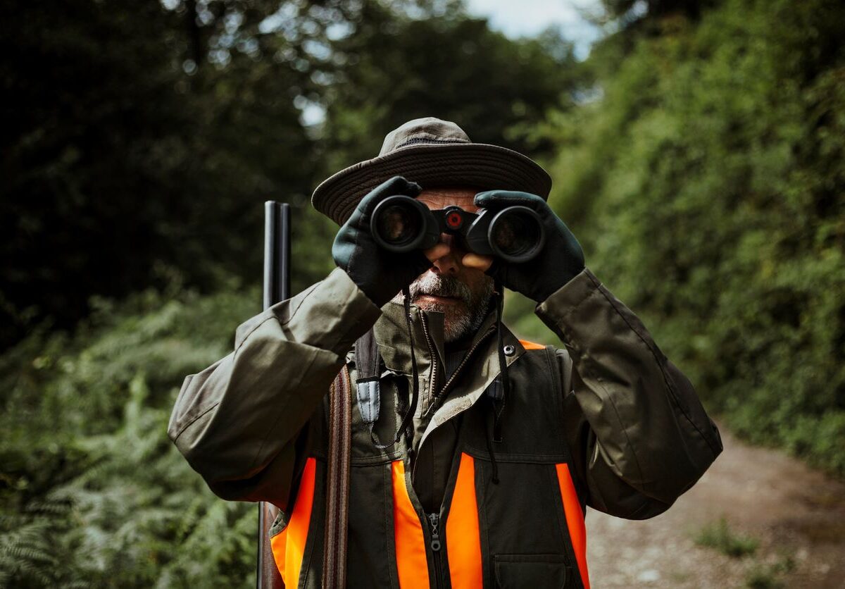 A man stands looking through his binoculars while hunting during the fall.