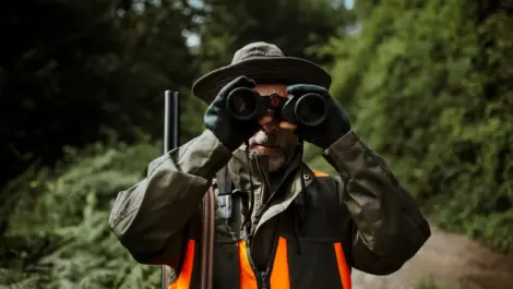 A man stands looking through his binoculars while hunting during the fall.