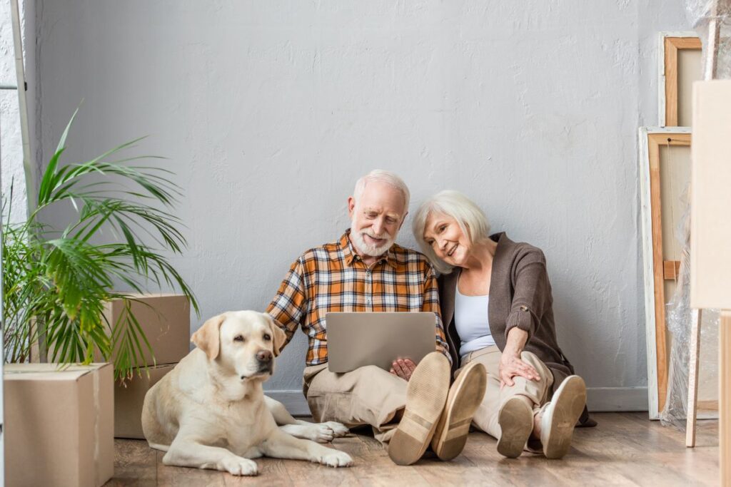 An older man and woman sitting with their dog next to boxes in a mostly empty apartment.