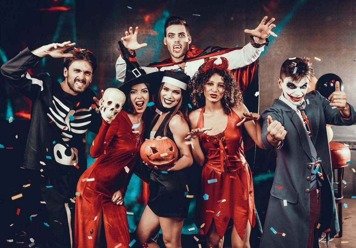 A group of young people at a Halloween party wearing spooky costumes, including a skeleton, vampire, devil, and Joker.