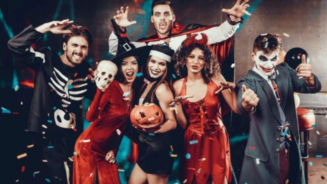 A group of young people at a Halloween party wearing spooky costumes, including a skeleton, vampire, devil, and Joker.