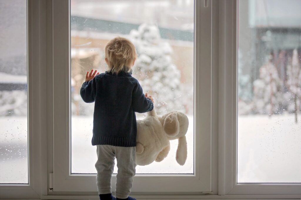 A boy holds his stuffed animal while looking out the window at his snowy yard.