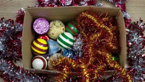 An open cardboard box filled with colorful Christmas ornaments and tinsel, showing mixed holiday decor stored loosely.